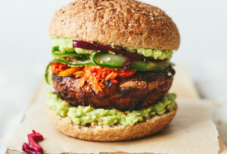 View Spicy miso mushroom burger One stacked spicy miso mushroom burger with all ingredients layered between a burger bun, displayed on a chopping board