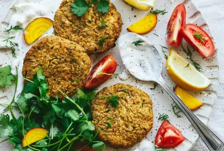 View Courgette & chickpea fritters with probiotic tzatziki One plate with three cooked courgette and chickpea fritters, served with probiotic tzatziki, fresh greens and tomato