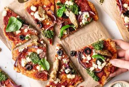 View Fibre-filled wholewheat & bean flatbread pizza A photo of flatbread pizzas on a wooden board. One slice is being grabbed.