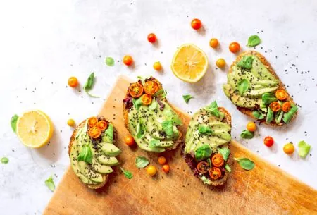 View Can an avocado a day keep high cholesterol away? 4 slices of toast topped with sliced avocado, fresh tomato and basil on a white surface and wooden chopping board with halved lemons and whole cherry tomatoes