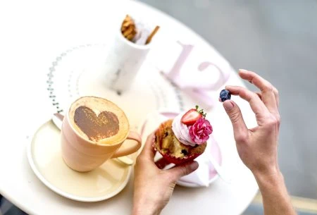 View Coffee & the gut A cup and saucer with coffee and chocolate love heart dusted on top with a cupcake topped with pink icing, a pink flower and a strawberry and blueberry picked off by a hand in the picture