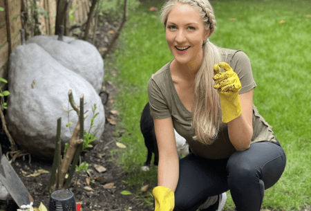View The hygiene hypothesis: what is it & what you can do Dr Megan Rossi squatting down in garden outside, wearing gloves with one hand in the soil