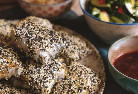 View Crispy plant protein-rich rice paper dumplings Rice paper dumplings covered in black and white sesame seeds on a plate with smaller plates in the background containing dips and salad