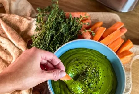 View Pistachio & white bean dip One small bowl full of pistachio and white bean dip. Carrot slices and fresh thyme accompanying, and a hand dipping into dip