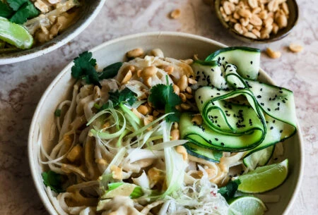 View Satay noodle bowl Satay noodles with ribbons of courgette, roasted peanuts, lime and sauerkraut and black sesame seeds with a bowl of peanuts in the background