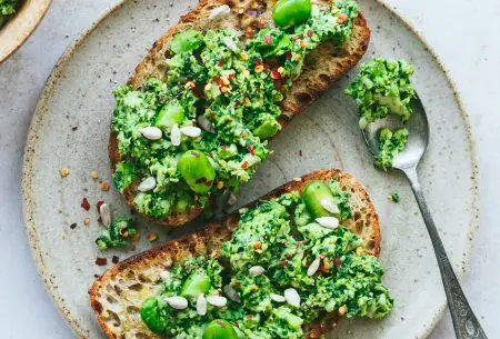 View Broad bean chilli toast Two pieces of toast on a plate, spread thick with broad bean chilli paste and mixed seeds