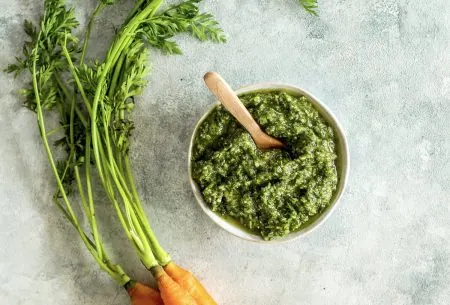View Anti-food waste carrot top pesto Pot of green carrot top pesto, shown on bench top with small bunch of fresh carrots and leafy green carrot tops