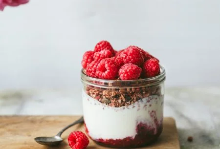 View Live yoghurt pots with raspberry compote One yogurt pot with creamy live yogurt and crunchy granola, topped with fresh raspberries and displayed on a chopping board