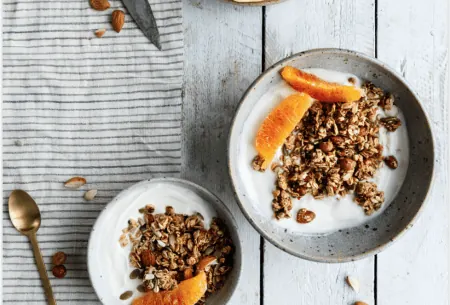 View Do you know what’s in your cereal? Two bowls of yogurt topped with granola and blood orange on a white wooden surface with a black and white striped tea towel, a spoon and a knife and a plate with segments of orange