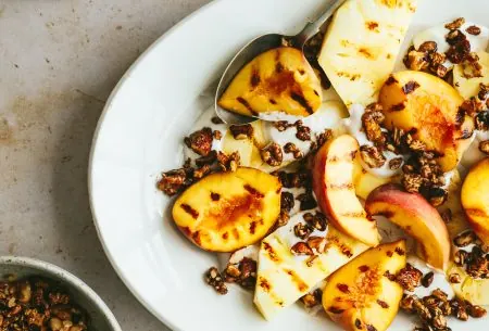 View Peaches & walnut crumble White plate with grilled peaches and walnut crumble, with an extra pot of walnuts to the side