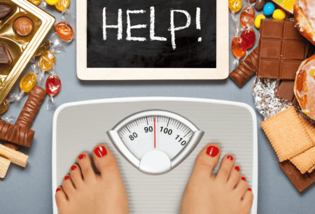 View Navigating your weight loss goals with binge eating disorder Two feet are shown standing on a set of scales with a sign reading 'help' in front, while background is covered by sweets.