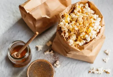 View Salt Awareness Week: how much is too much? A brown paper bag of popcorn and a cinnamon shaker and a glass pot of honey with a honey spoon
