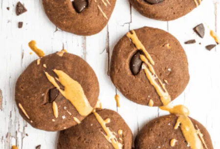 View Chocolate & peanut butter teff cookies Six chocolate & peanut butter teff cookies on bench top with chocolate chunks and peanut butter drizzle