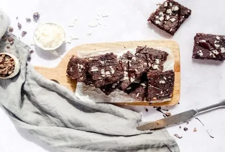 View Chocolate & coconut brownies Eight chocolate and coconut brownies stacked along a chopping board, topped with coconut flakes