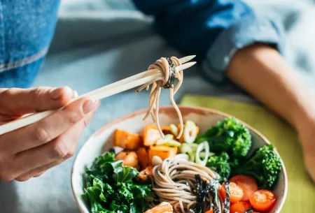 View The key to gut health: plant diversity! A bowl of noodles, veggies and prawns with someone wrapping the noodles around chopsticks in a denim shirt