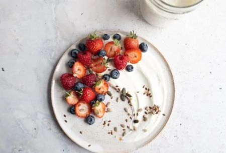 View Live yoghurt benefits A glass pot of yogurt alongside two plates with yogurt, seeds and berries